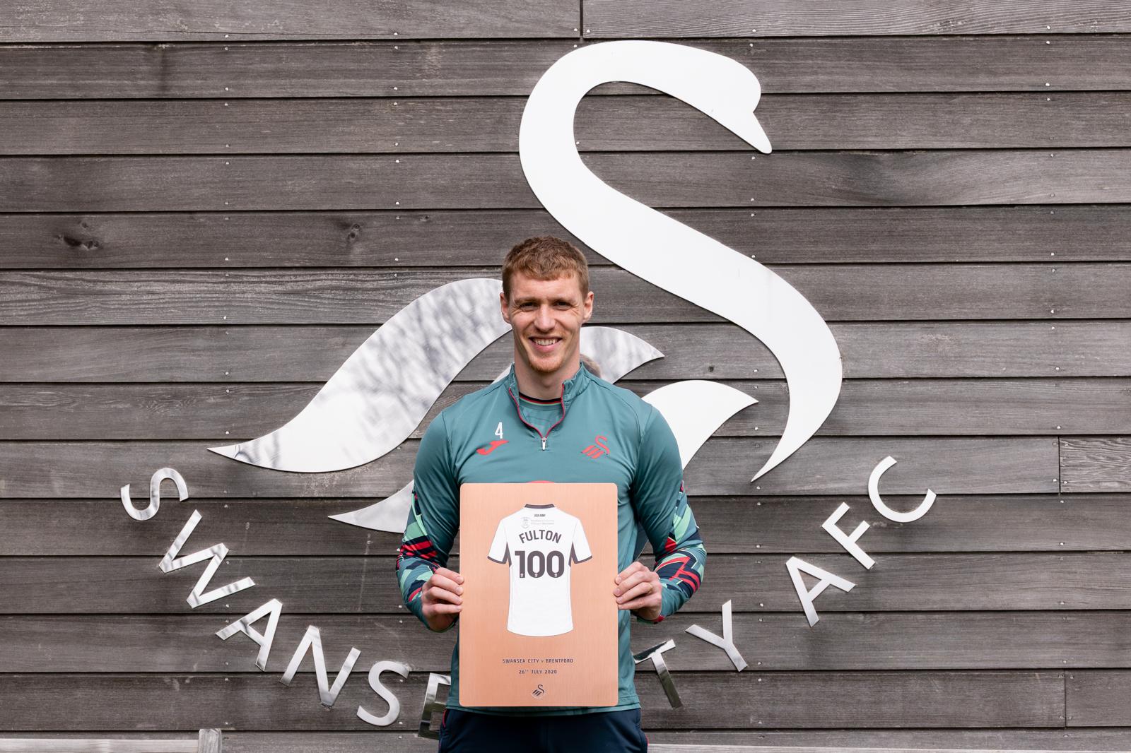 Jay Fulton holding his 100 appearance Copper finish plaque in front of the Swansea City AFC swan logo