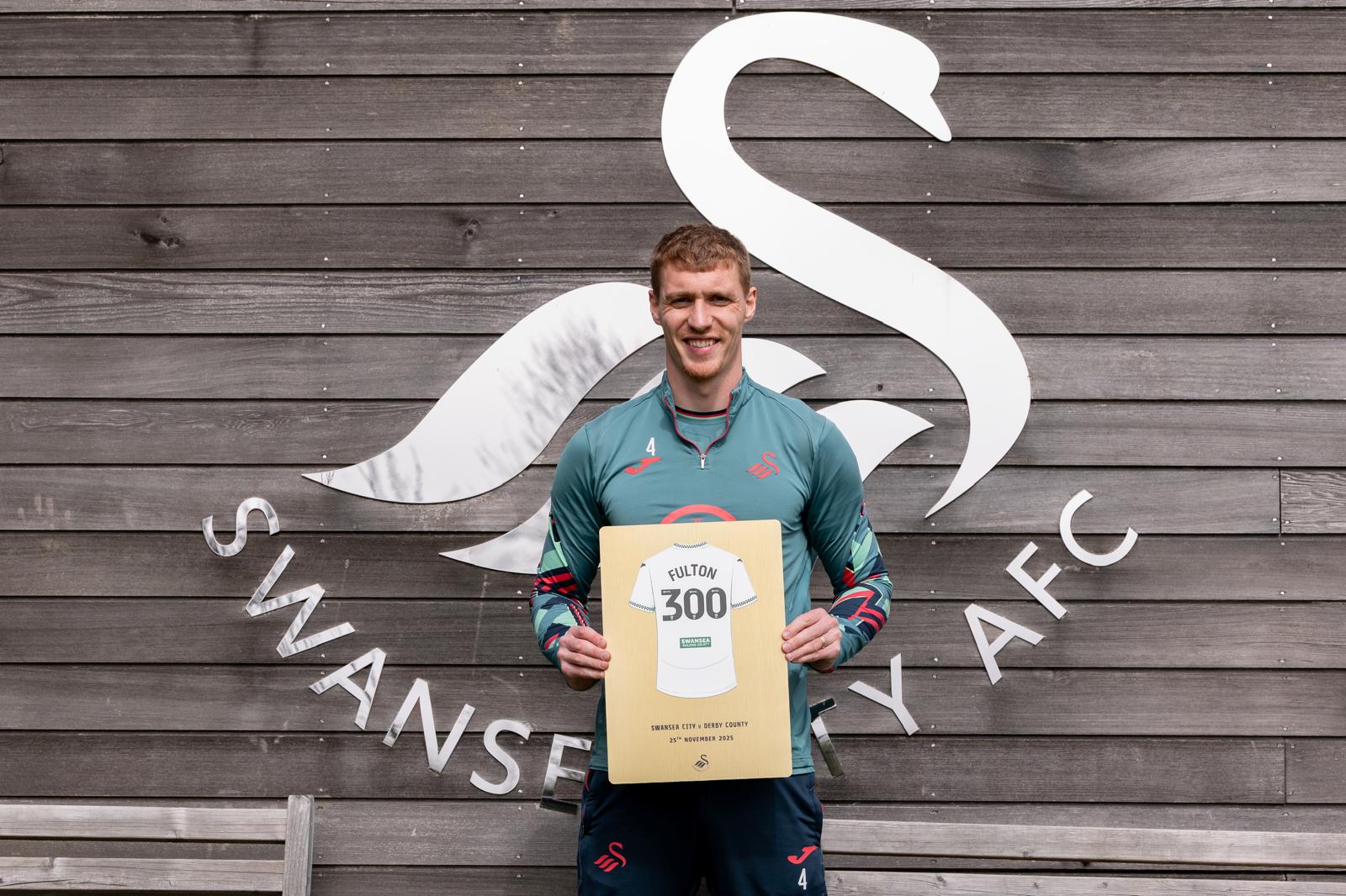Jay Fulton holding his 300 appearance Gold finish plaque in front of the Swansea City AFC swan logo